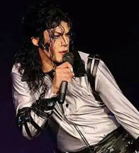 Michael Jackson african american male with wavy shoulder length black hair wearing a grey collared button up shirt with black leather stripes on the shoulders.