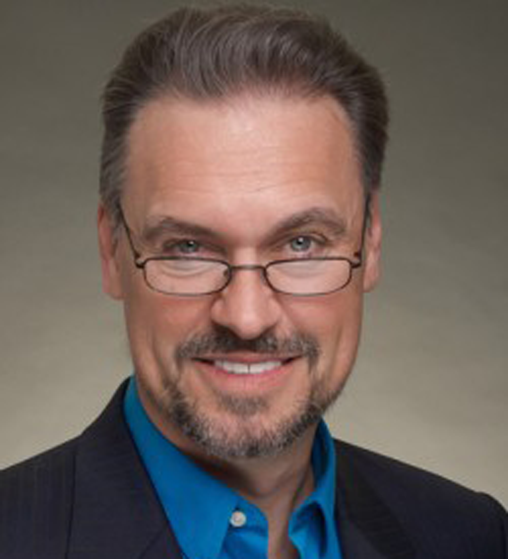 Ron Smith caucasian male with grey hair and a clean cut goatee, wearing black thin rimmed prescription glasses, a black suite with a blue collared shirt.
