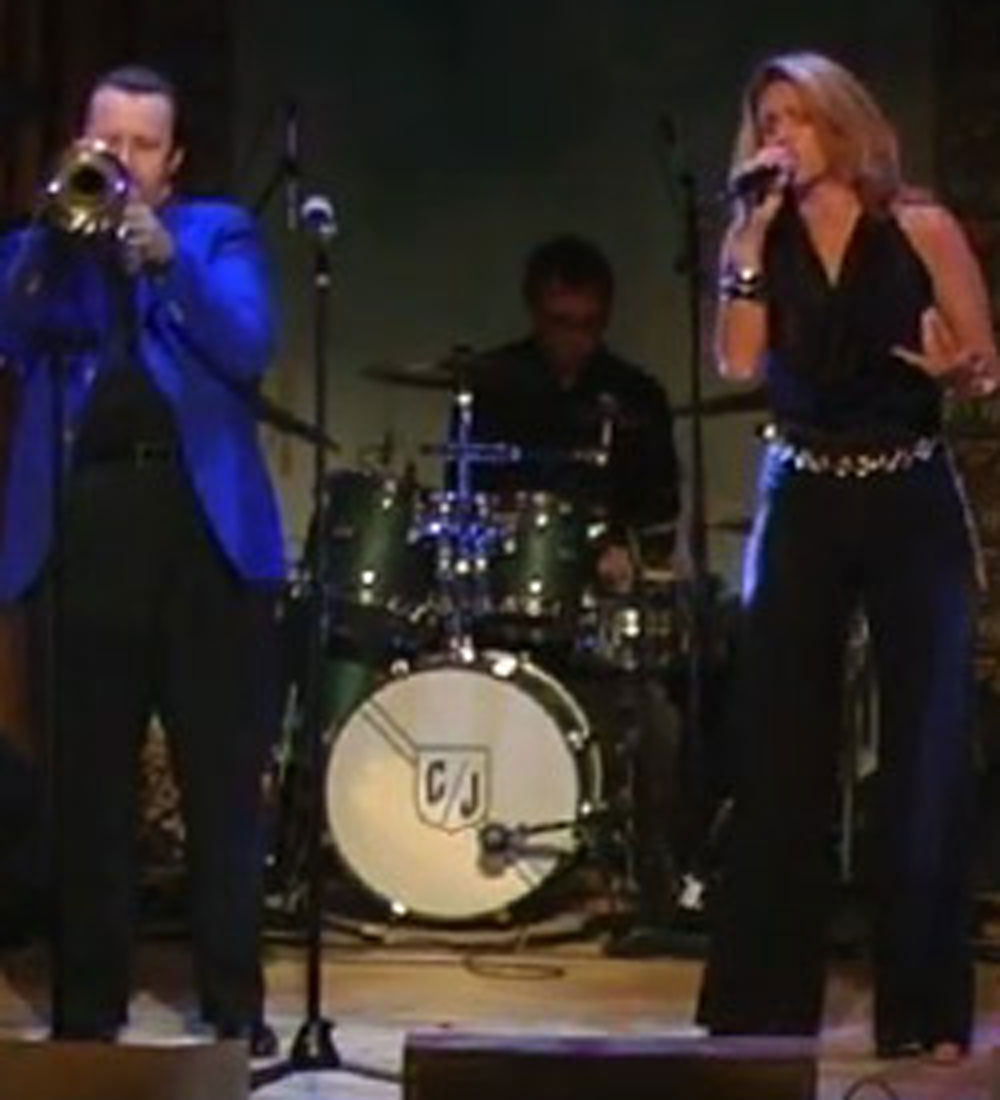 Bands cover photo with one caucasian male wearing a royal blue blazer and a caucasian woman wearing an all black romper and a drummer in the back wearing an all black hat and a black shirt.