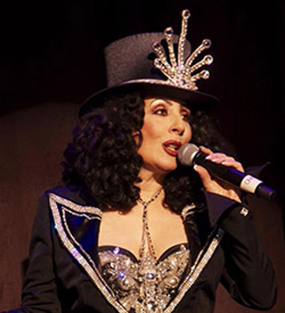 Cher celebrity impersonator caucasian woman with curly black hair wearing a black top hat with a diamond sculpture design in the front and wearing a black blazer trimmed in diamonds and a diamond embezzled corset.