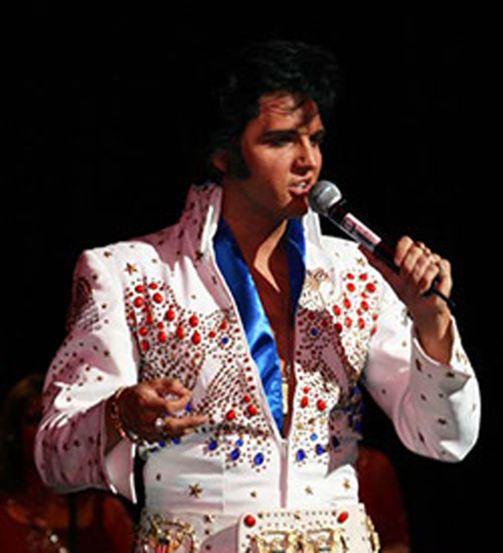Elvis celebrity impersonator caucasian male with short black hair wearing an all white jump suit dazzled with colorful jewels on the front.